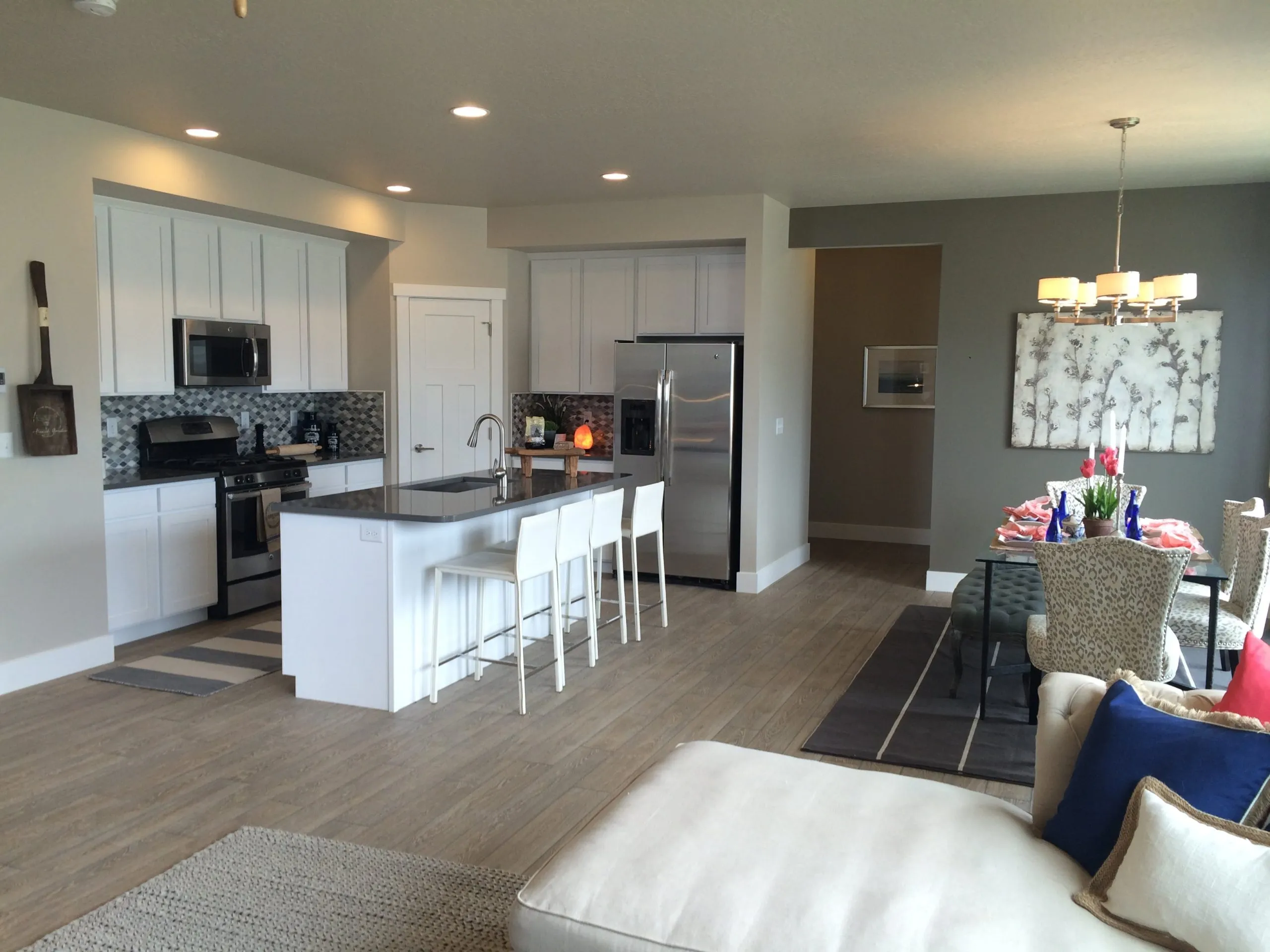 Modern open-concept kitchen and dining area found in many homes in Utah, featuring white cabinets, stainless steel appliances, a kitchen island with three white stools, and a dining table for six by a window with a chandelier above.