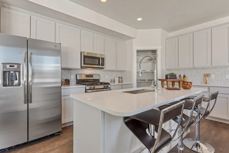 Modern kitchen with white cabinets, stainless steel appliances, and a center island with a sink—perfect for homes in Utah. Three dark barstools complement the tiled backsplash, while utensils and decor add charm to the counters.