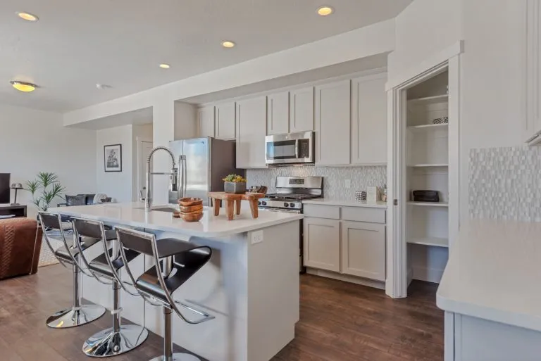 Modern kitchen with white cabinets, stainless steel appliances, a center island with bar stools, light countertops, open pantry, wood flooring, and decorative plants—a perfect example of bright and open homes in Utah.