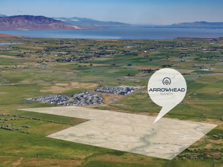 Aerial view of green fields, a residential area featuring homes in Utah, distant mountains, and a shaded area labeled Arrowhead Ranch marked by a pin. A lake can be seen in the background.