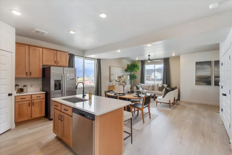 Modern open-concept kitchen and living room with light wood floors, wood cabinets, stainless steel appliances, island with sink, dining table, large windows, and neutral décor with plants and artwork.