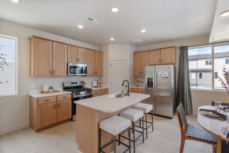 Modern kitchen with light wood cabinets, stainless steel appliances, a center island with three cushioned stools, and a dining table. Large windows let in natural light, creating a bright and airy space.