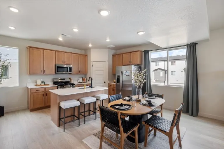 Modern kitchen and dining area with wooden cabinets, stainless steel appliances, a center island with bar stools, and a dining table set for four near large windows with gray curtains. Natural light fills the space.