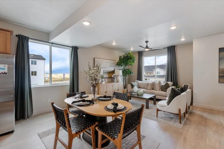 A modern open-concept living and dining area features a round table set for four, woven chairs, a cozy sofa, large windows with dark curtains, light wood floors, and contemporary decor.