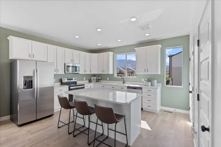 Modern kitchen with white cabinets, stainless steel appliances, a center island with three brown chairs, light wood floors, and large windows letting in natural light and showing a mountain view outside.