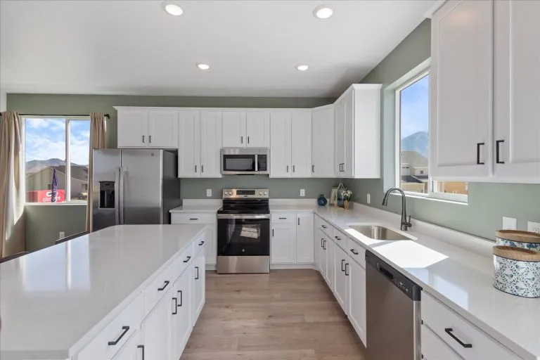 Modern kitchen with white cabinets, stainless steel appliances, light wood flooring, large island, and green walls. Sunlight streams in through two windows, creating a bright, airy atmosphere.