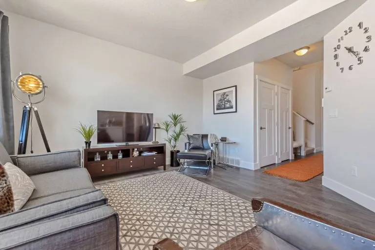 Modern living room with gray sofa, TV on a wooden stand, potted plants, tripod lamp, and patterned rug—features often found in stylish homes in Utah. White walls and doors, hardwood floors, and a staircase complete the inviting space.
