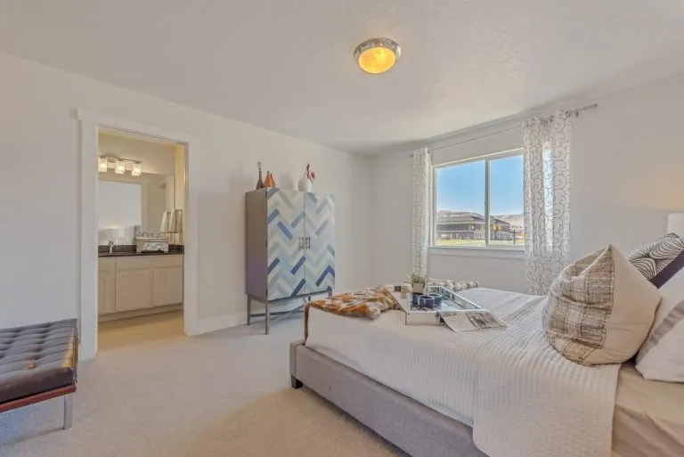 A bright, modern bedroom in homes in Utah features a large bed, patterned cabinet, bench, and open doorway to a well-lit bathroom. Sunlight streams through sheer curtains, highlighting the neutral decor and inviting atmosphere.