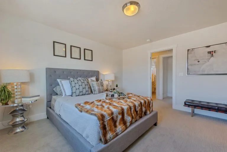 Modern bedroom with a gray upholstered bed, patterned pillows, fur throw, and wall art—showcasing the elegance found in many homes in Utah; an open door reveals a glimpse of a well-lit bathroom.