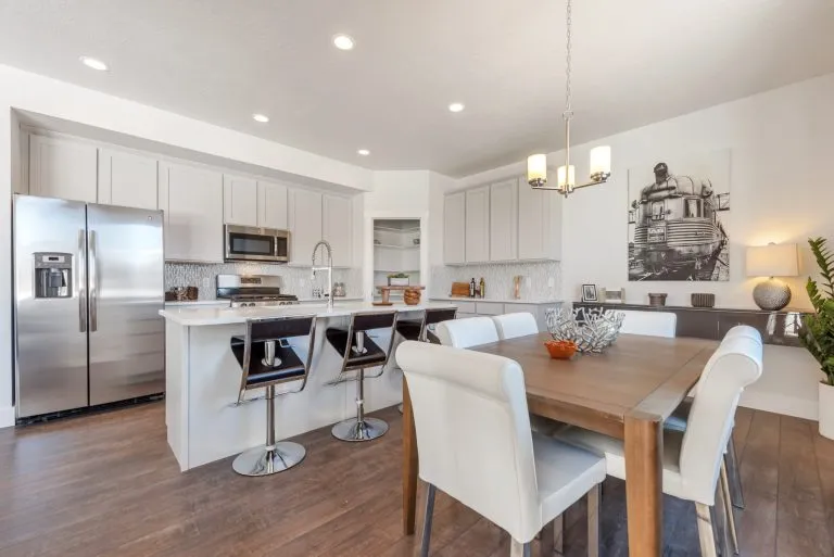 Modern kitchen and dining area found in many homes in Utah, featuring white cabinets, stainless steel appliances, a kitchen island with black barstools, wooden dining table with white chairs, and a decorative photo of a train on the wall.