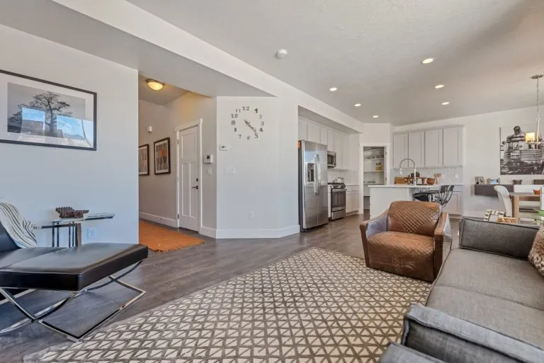 Modern open-concept living room and kitchen in homes in Utah with hardwood floors, neutral furniture, geometric rug, wall clock, stainless steel appliances, and white cabinets. Bright with natural and recessed lighting.