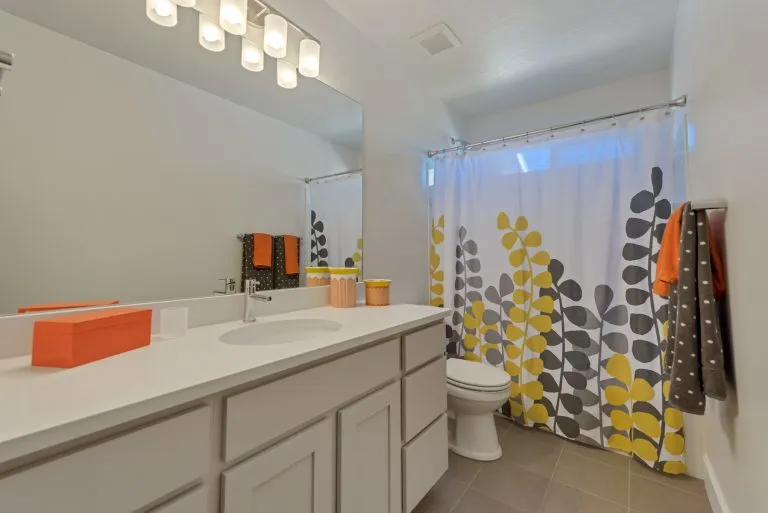 Bright bathroom with white cabinets, a large mirror, and modern lighting above—common features in many homes in Utah. The shower curtain’s yellow and gray leaf pattern pairs nicely with orange and gray towels, adding pops of color to the neutral-toned space.