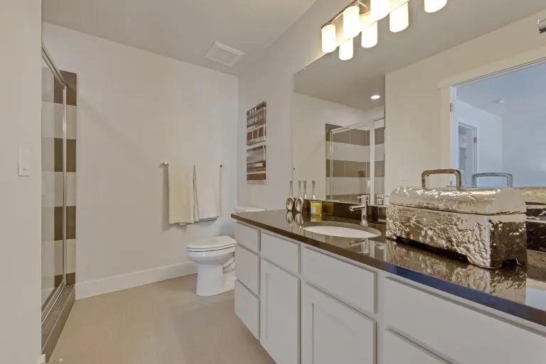Modern bathroom in homes in Utah featuring a glass shower, white cabinets, black countertop, large mirror, towel rack with folded towels, and decorative items near the sink under bright vanity lights.
