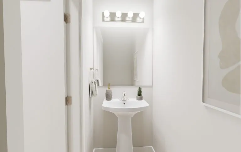 A small, modern bathroom in homes in Utah features white walls, a pedestal sink, a large mirror with a row of lights above. The sink holds soap, a toothbrush holder, and a small potted plant. Towels hang on the wall to the left.