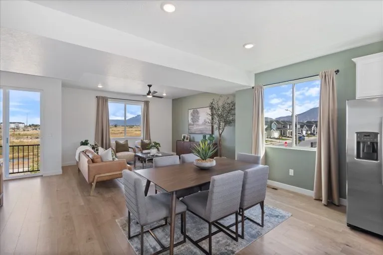Modern open-concept living and dining area with large windows, mountain views, a wooden dining table with six gray chairs, a light sofa, and contemporary decor in neutral tones.