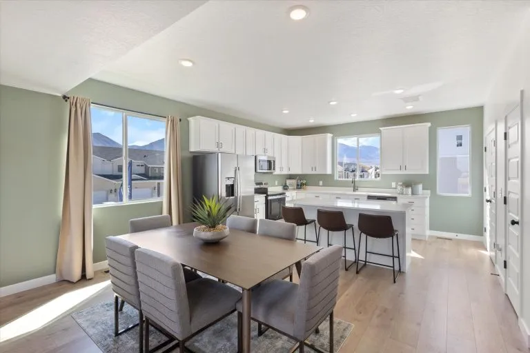 Modern kitchen and dining area with light green walls, white cabinets, stainless steel appliances, a wooden dining table with six upholstered chairs, a kitchen island with three barstools, large windows, and wood flooring.