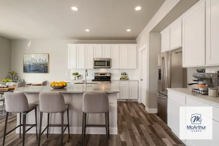 Modern kitchen with white cabinets, stainless steel appliances, a kitchen island with three gray barstools, and wood-style flooring. A dining area and artwork complete this inviting space in homes in Utah. McArthur Homes logo is in the corner.