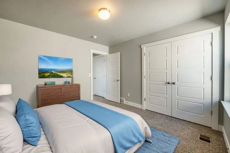 A modern bedroom in homes in Utah, featuring a neatly made bed with blue and white bedding, a wooden dresser topped by a landscape photo, double white closet doors, beige carpet, and light gray walls.