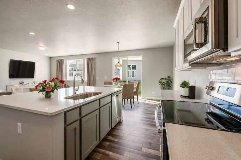 Modern kitchen with white cabinets, stainless steel appliances, and an island with sink—perfect for homes in Utah. Fresh flowers adorn the island and dining table, while large windows open into a bright living and dining area.