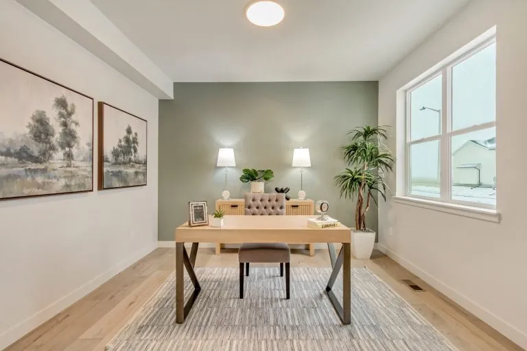 A modern home office with a wooden desk, cushioned chair, large window, indoor plant, two lamps, decorative art on the wall, and soft neutral tones throughout the room.