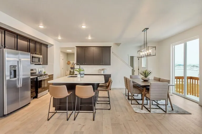 Modern kitchen and dining area with dark cabinets, stainless steel appliances, island with sink and three barstools, dining table with six chairs, large windows, and light wood flooring.