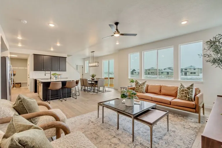 Modern open-concept living room and kitchen with neutral colors, large windows, a tan sofa, armchairs, a coffee table, barstools at the island, dining area, and ceiling fan. Natural light fills the space.