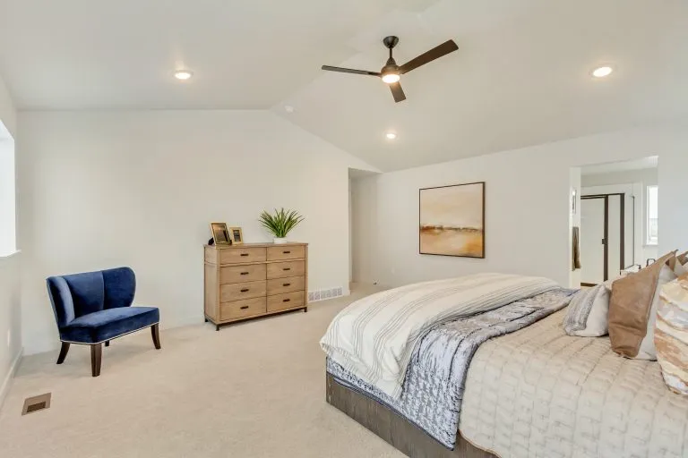 Spacious bedroom with a neatly made bed, light walls, beige carpet, a wooden dresser with decor, a blue accent chair, modern ceiling fan, and a large abstract painting on the wall.