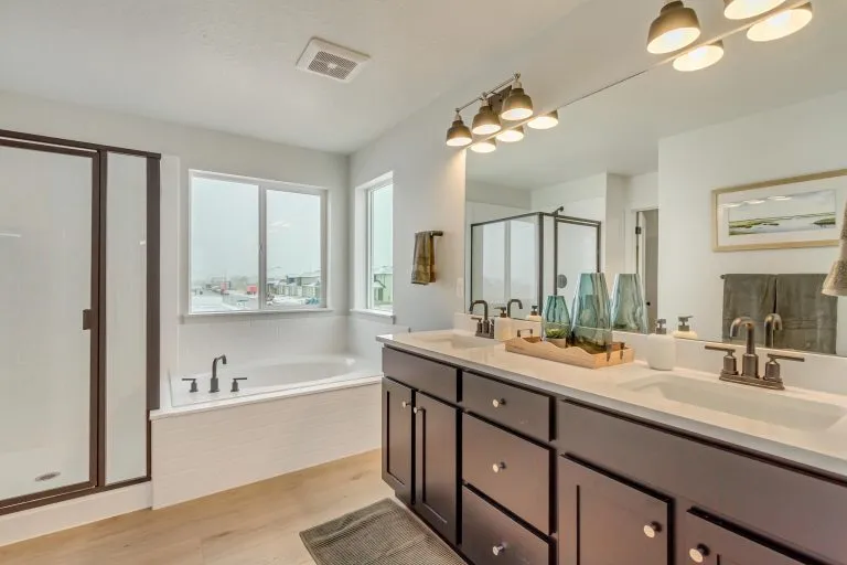 Modern bathroom with a dark wood double-sink vanity, large mirror, light fixtures, glass-enclosed shower, and a bathtub beneath two windows. Decorative vases sit on the counter. Walls are light colored with wood flooring.