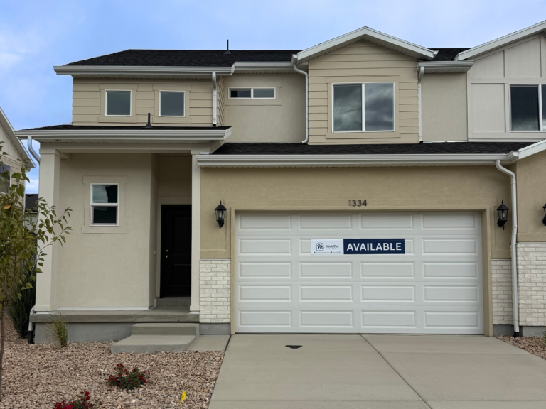 Two-story townhome with tan siding, dark roof, single-car garage, and an Available sign posted near the front door.