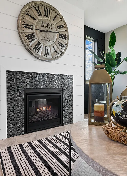 A cozy living room features a fireplace with a mosaic tile surround, a large vintage-style wall clock above it, a striped rug, a table with candles, a globe, and a green plant by the window.