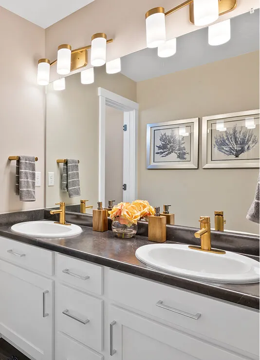 Double bathroom vanity with two white sinks, gold faucets, and matching soap dispensers. A vase of yellow flowers sits between the sinks. Above are modern gold and white light fixtures and a large mirror. Two framed prints hang on the wall.