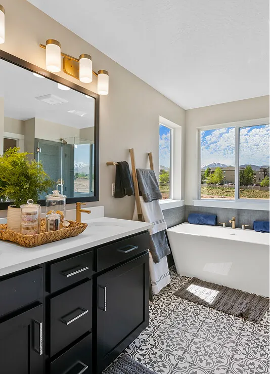 Modern bathroom with black vanity and gold fixtures, patterned tile floor, freestanding white tub by large windows, decorative ladder with towels, and a basket of toiletries on the counter.