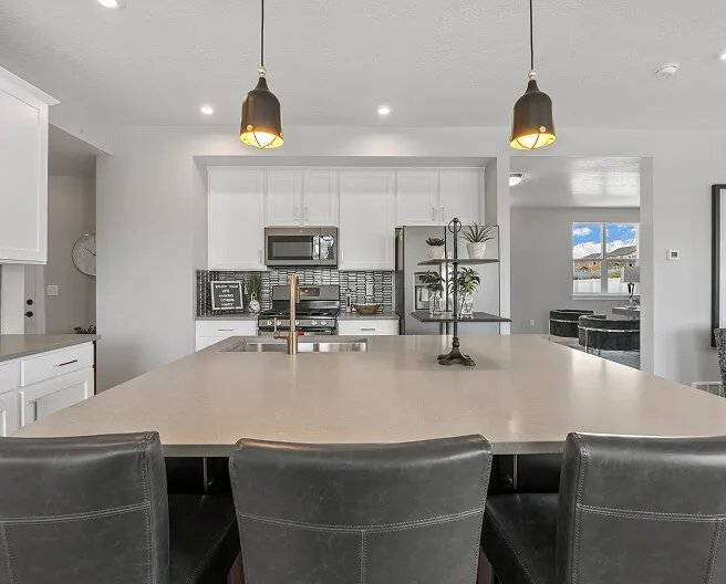 Modern kitchen with a large island, three black leather chairs, white cabinets, stainless steel appliances, pendant lights, and a window showing a view outside.
