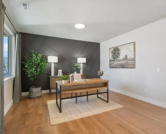 Modern home office with a wooden desk, laptop, and decorative items. Behind the desk are two floor lamps, a potted plant, and a large painting on the wall. The room has wood flooring and a large window with gray curtains.