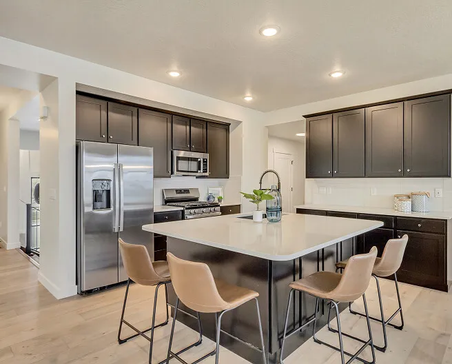 Modern kitchen with dark cabinets, stainless steel appliances, a large island with a light countertop, four tan barstools, and light wood flooring. Recessed ceiling lights brighten the space.