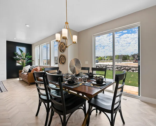 A modern dining room with a wooden table set for six, black chairs, a gold chandelier, large windows with a view of a green lawn and rocks, light wood floor, and decorative wall art. A sunlit sitting area is in the background.