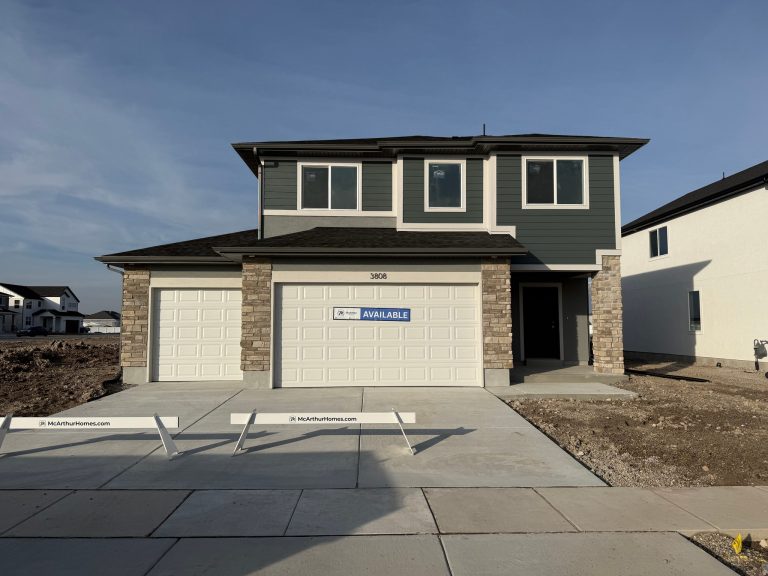 Two-story McArthur home with green and gray siding, stone accents, three-car garage with white doors, and an Available sign in a new development.