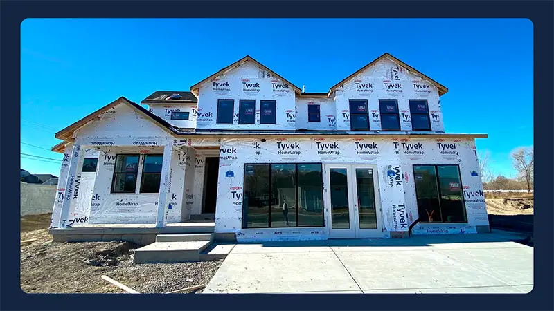 Two-story home under construction with Tyvek house wrap, windows installed, and concrete patio poured on a clear day