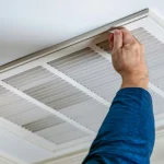 Person in blue shirt removing a white ceiling air return vent cover to access and replace the furnace filter inside.
