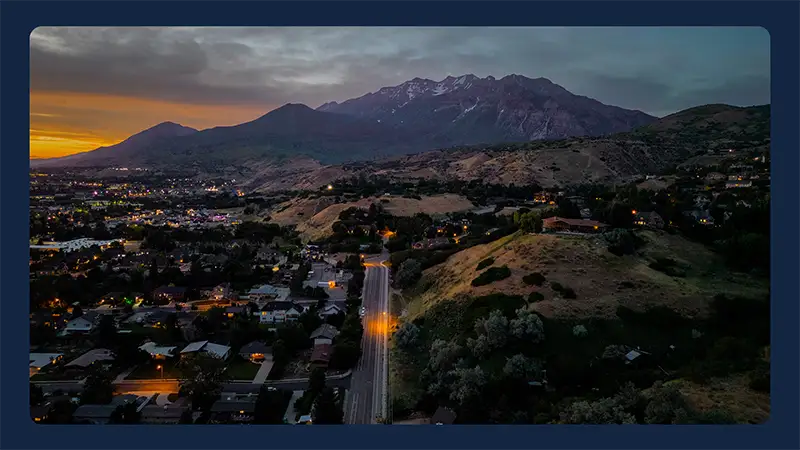 Aerial view of a Utah neighborhood at sunset with mountain peaks and glowing streetlights along a main road