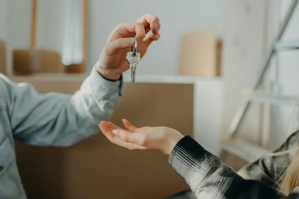 A person hands a set of house keys to an outstretched palm in front of stacked cardboard moving boxes inside a new home.